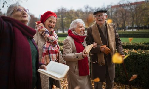 Group of happy senior friends with books on walk outdoors in park in autumn, talking and laughing